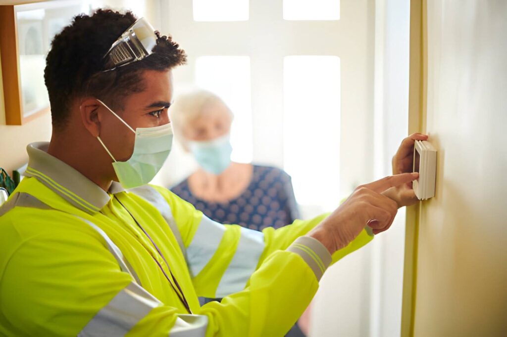 A masked technician in a high-vis vest adjusts a smart thermostat on a wall while an elderly woman watches